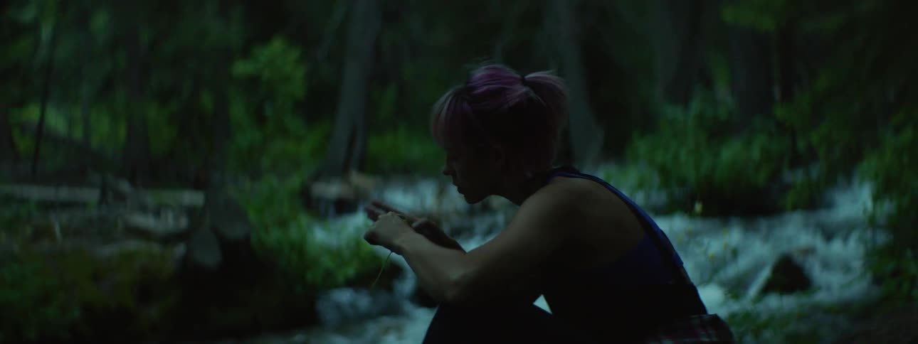 A young woman sits by the side of a rushing river while inspecting a small wild flower