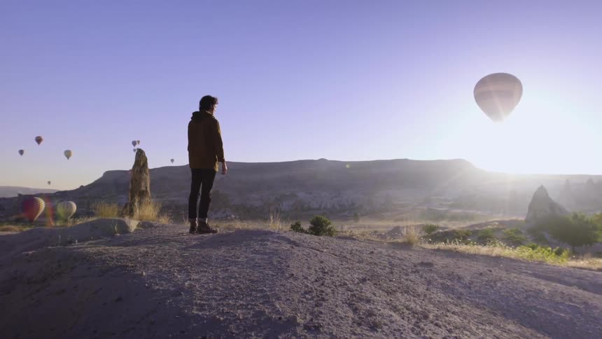 A man is standing on a hill looking at hot air balloons in the sky