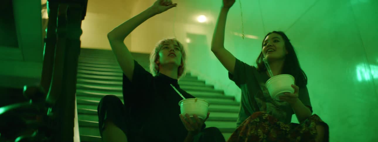 Two women sitting on a set of stairs playfully eat individual noodles from bowls while smiling