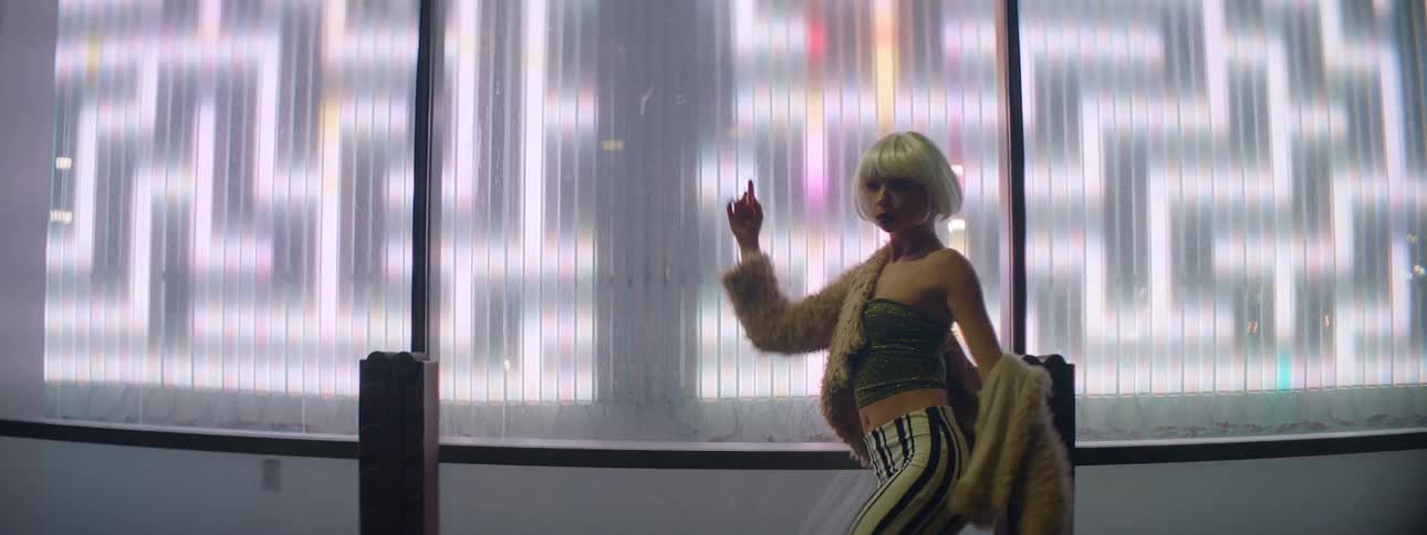 A woman wearing a blonde wig dances in front of an electronic screen that changes from a maze to a downtown city at night