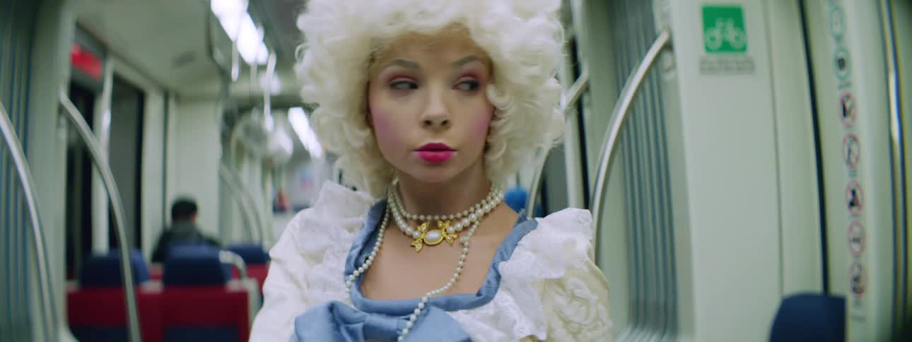 A young woman in an old fashioned dress and puff white wig dances while standing in the aisle of a train