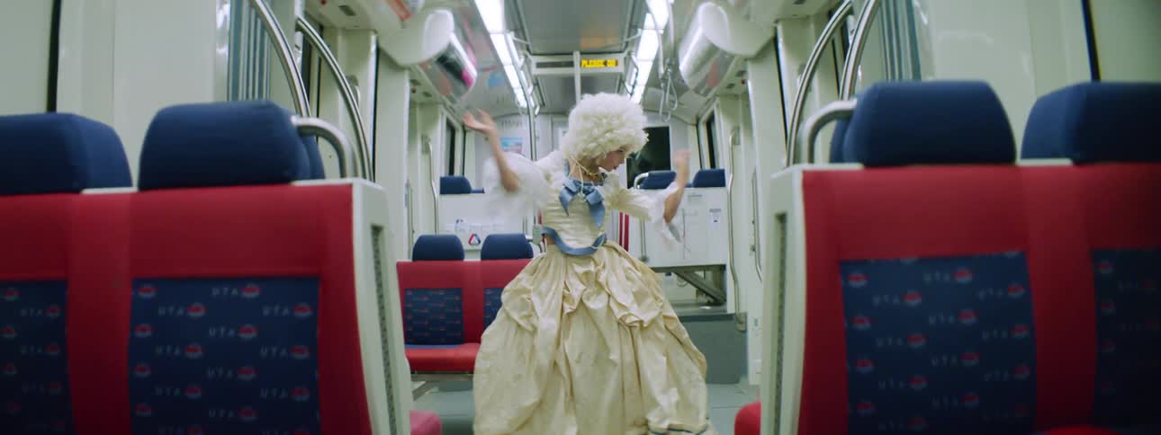 A woman in old fashioned clothing and a wig dances in the aisle of an empty bus as it travels at night