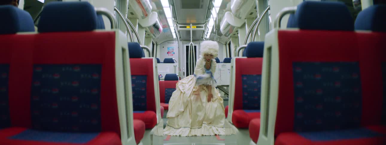 A woman in an old fashioned dress and powdered wig dances in a moving subway car