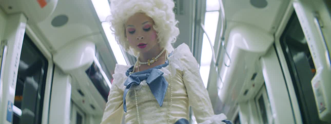 A woman in a victorian era dress and a powdered wig dances in the middle of a subway car