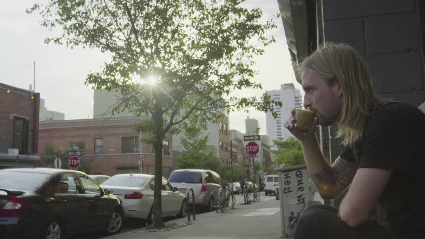 A man with long blonde hair sits outside in the city drinking from a small mug