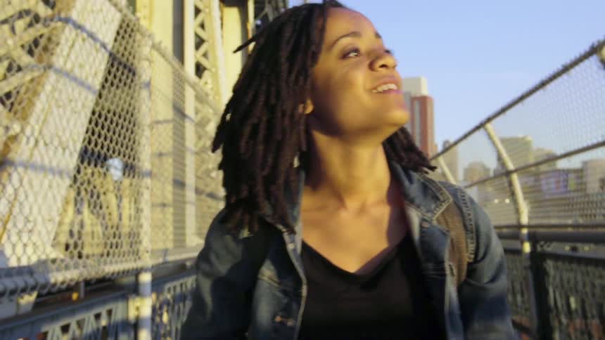 A smiling woman walks across a fenced bridge in the city on a sunny day