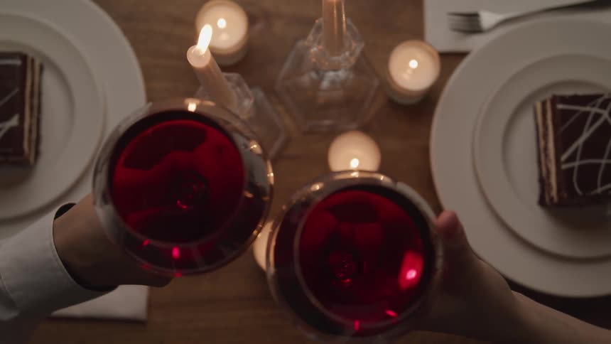 Two people celebrate with red wine while dining at a table with candles