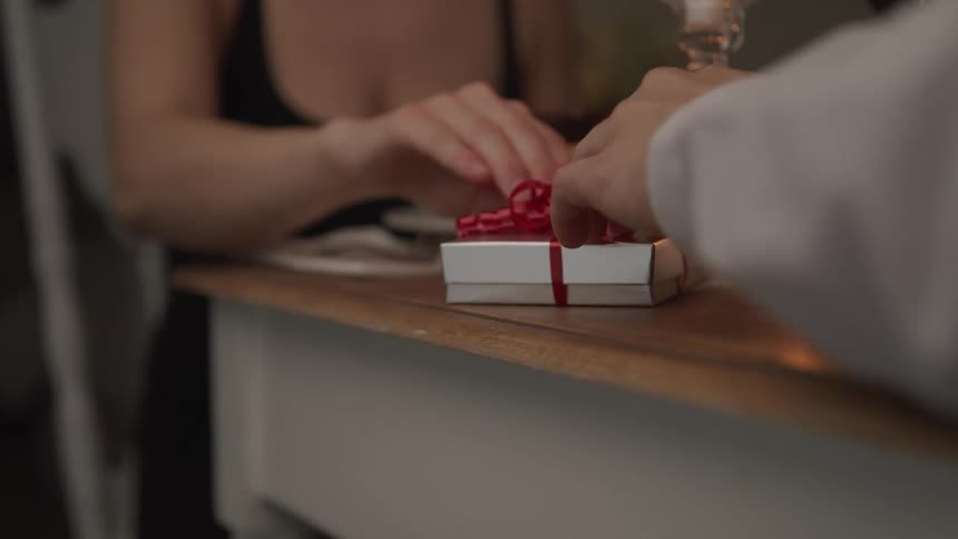 Male pushes a wrapped gift forward to female on the other side of the candle lit table as she reaches forward to accept it