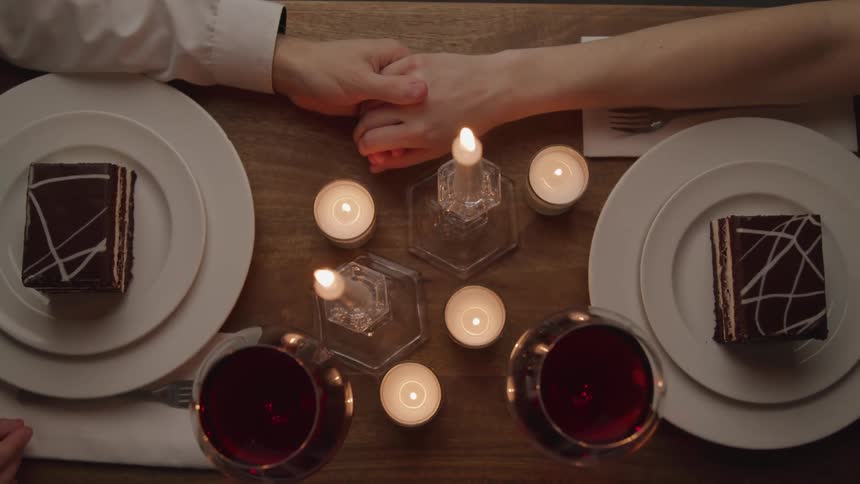 A man and woman reach across a table with candles wine and dessert to hold hands