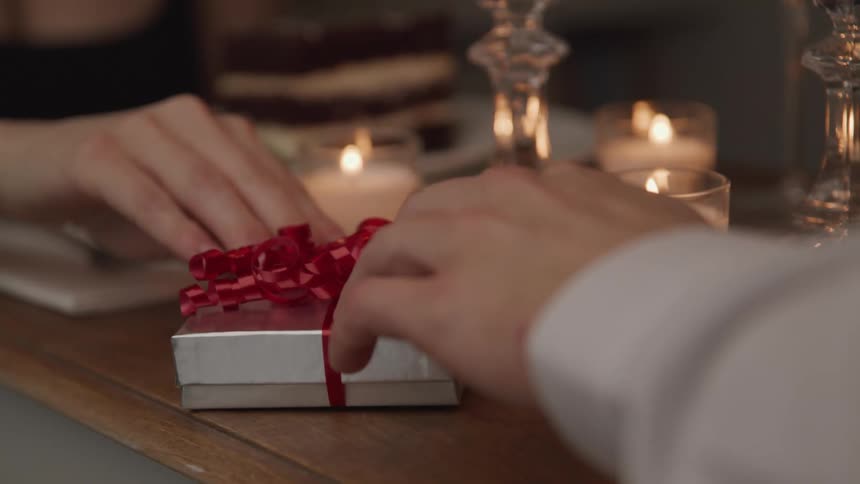 A person uses their hand to slide a small gift box wrapped in red ribbon across a candlelit table to the person sitting on the other side