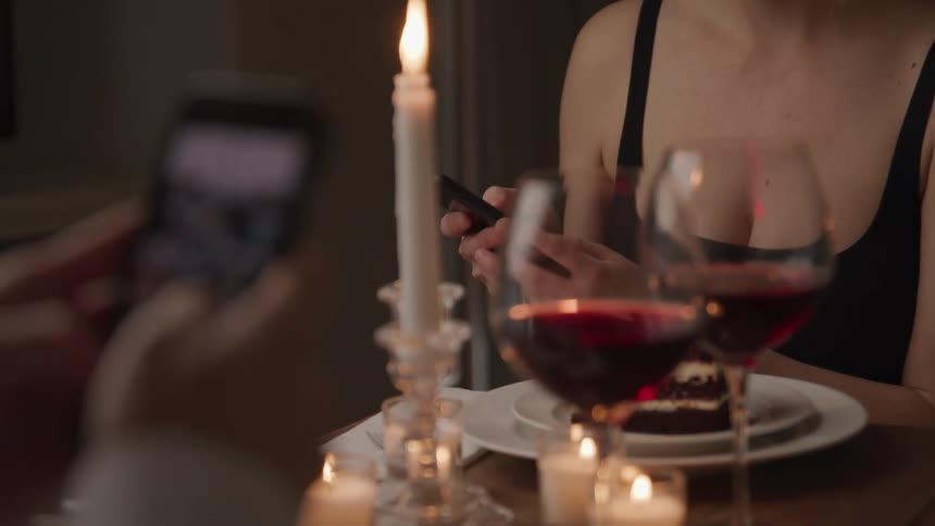 Two people sitting at table for candlelight dinner both use their mobile phone while dessert waits on plate