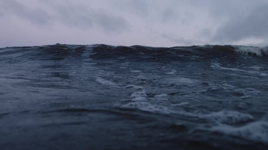 The water is rough and a wave crashes under a cloudy sky