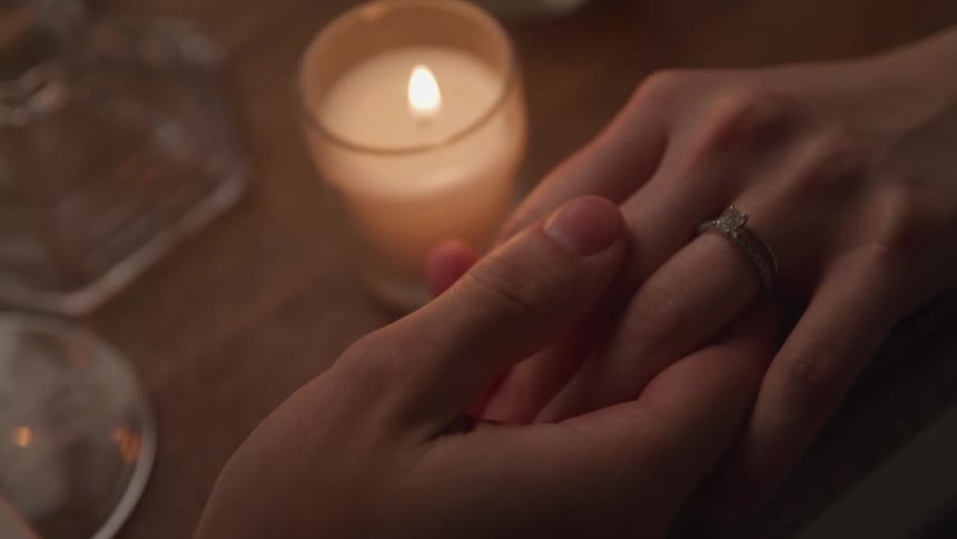 Male holds females hand across a candle lit table while caressing the top of her fingers with his thumb