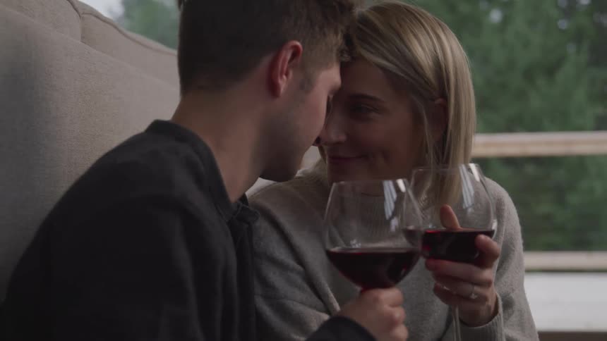 A man and a woman sitting holding wine glasses with wine as their foreheads touch