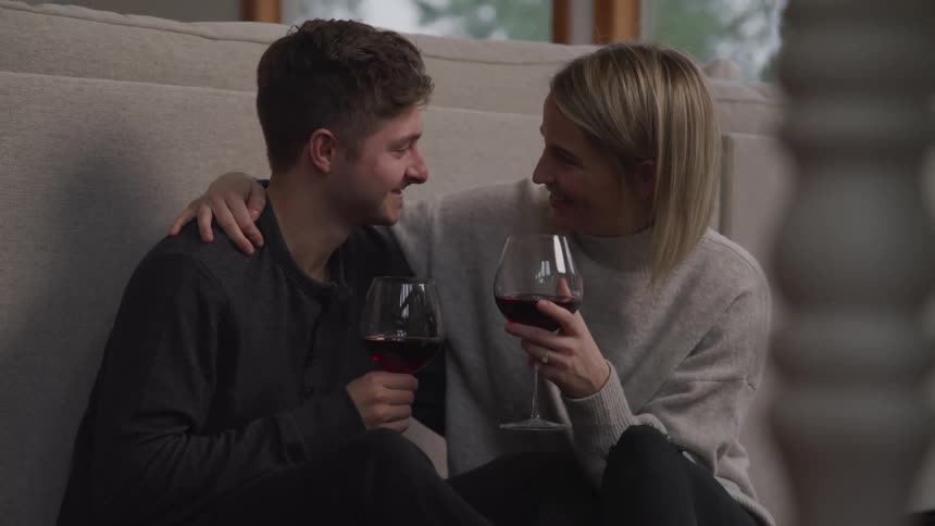 A woman has her arm around her husband as they sit on the floor holding wine glasses