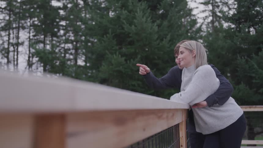 A man and a woman standing on a porch hugging with trees and the sky in the background