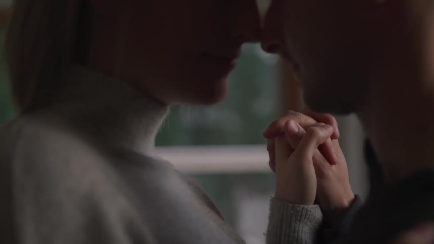 A man and woman stand with their noses pressed together as they hold hands in a house