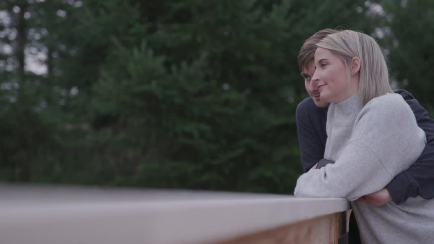 A man and woman hug while leaning against a deck railing surrounded by trees