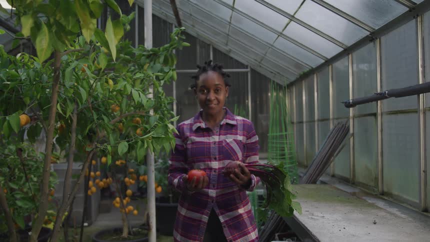 A woman stands in a green and smiles as she holds a tomato in her left hand and a beet in the other