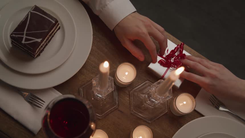 A man passes a small gift box across a wooden dining table to a woman sitting opposite him