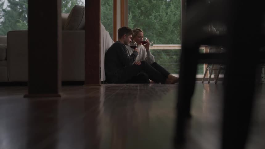 Young couple sits in the floor of the living room drinking wine reclined on a couch back
