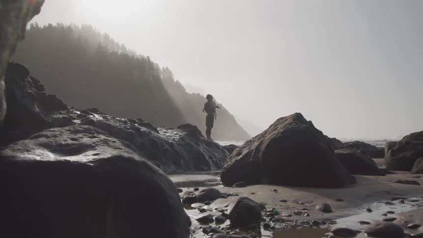 A man standing on a large rock on the shore stretches his arms up towards the sky