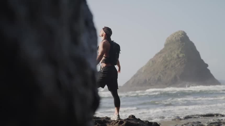 Shirtless fit man wearing a backpack stands on a rock in the beach next to the ocean