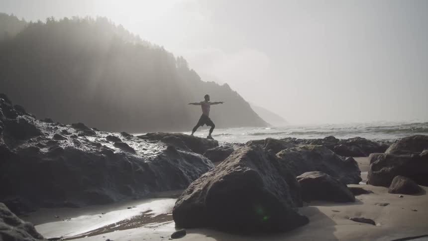 A shirtless man does yoga atop large rocks on a beach below a mountain shrouded in fog