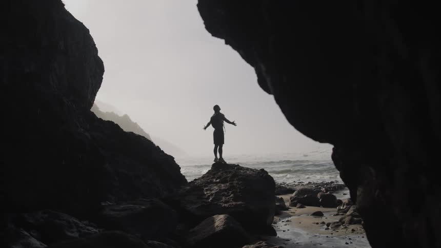 A young man stands on a rock on the shore and stretches his arms over his head