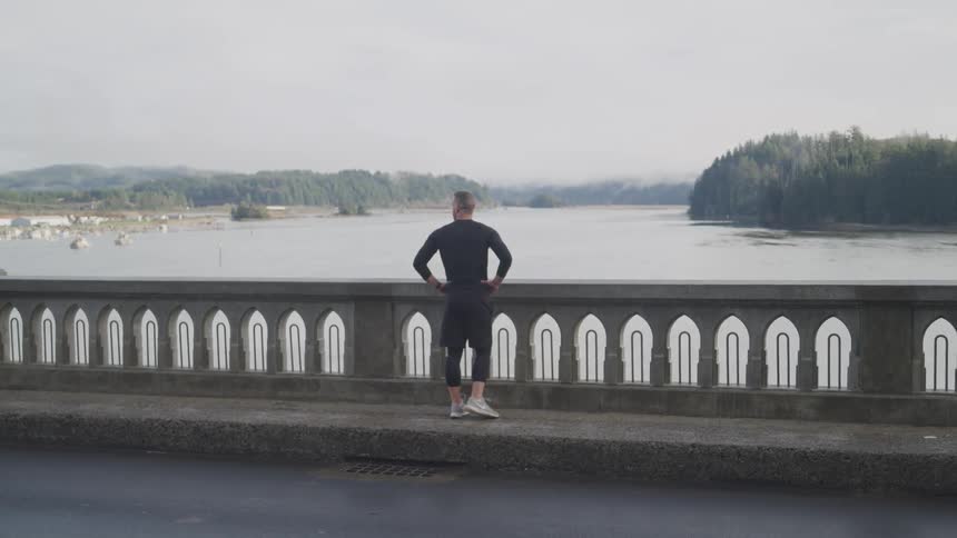 A man takes a break from jogging to stand and admire the river below from the bridge that he is standing on