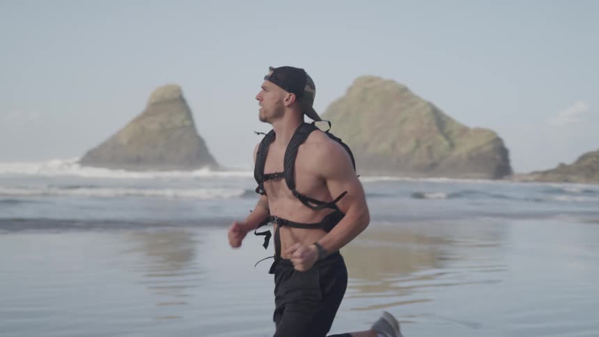 Fit shirtless young man wearing a backpack runs on the wet sand beach at sunset