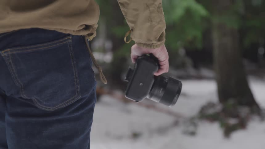 A man walks through a wooded area covered in snow carrying a camera