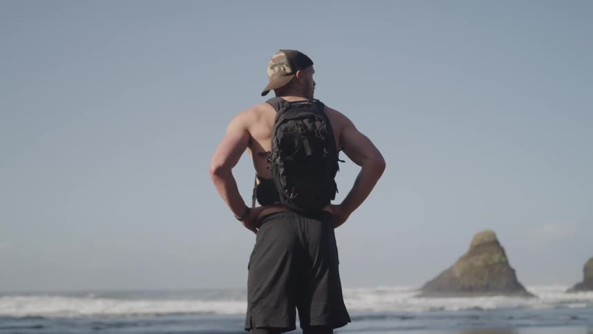 A shirtless man wearing a backpack stands on a beach and looks out over the ocean on a sunny day