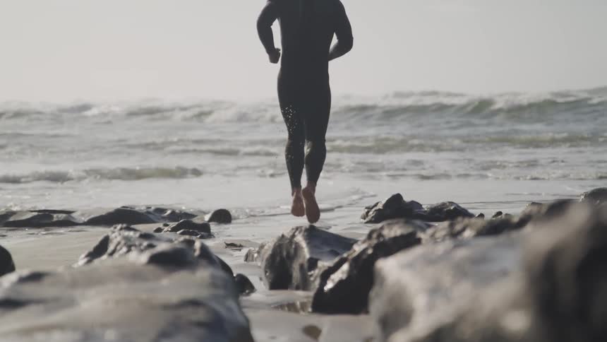 Young fit man wearing a black wet suit runs to the ocean on a beach