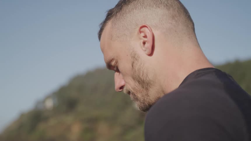 A man with his head tilted down and his eyes closed stands outdoors on a sunny day