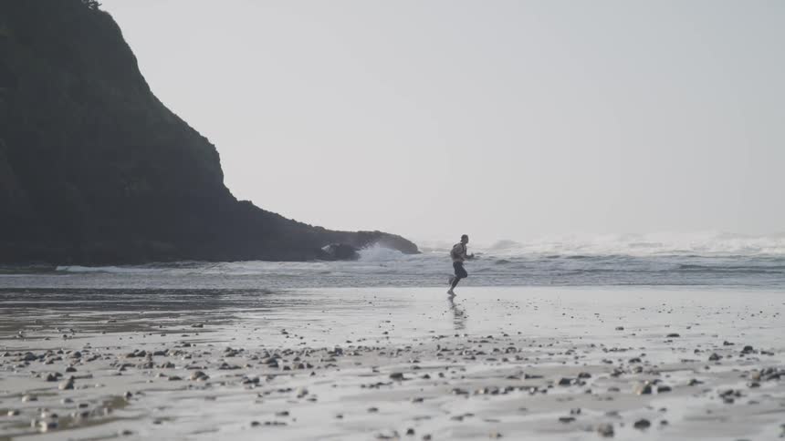 A shirtless man runs across a rocky beach under a cloudy sky