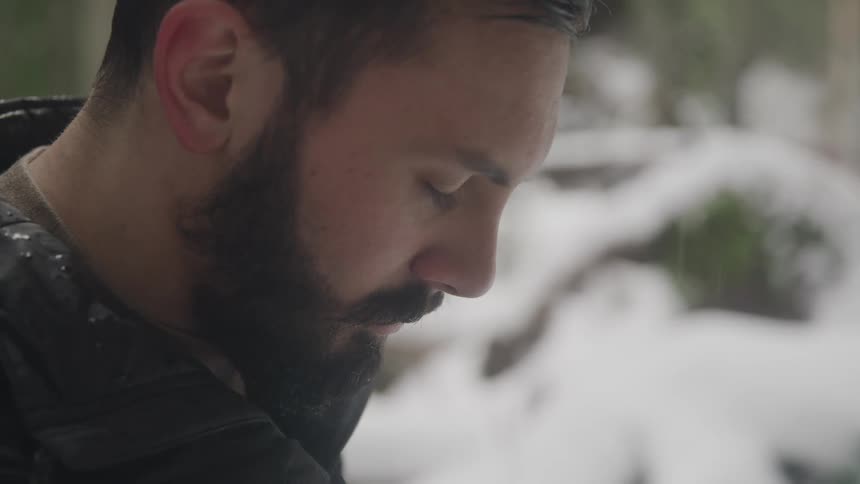 Side view of a man with a beard sitting on a tree trunk with eyes closed in the snowy forest