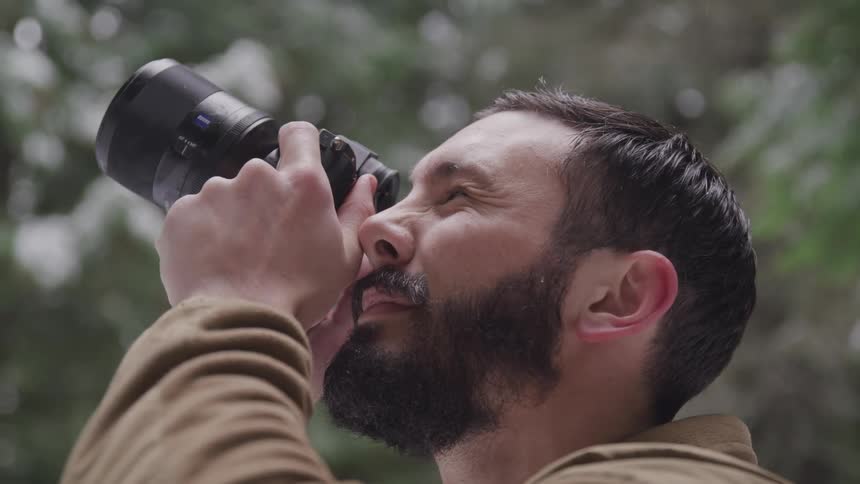 A bearded man raises a camera to his eye as he aims to take a photograph