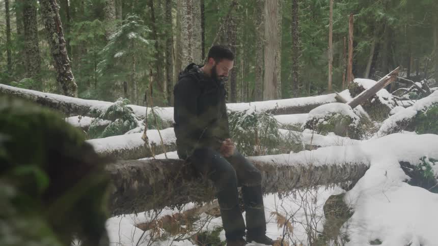 Man with a beard sits on a tree trunk in the snowy forest