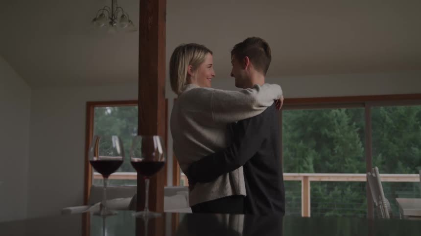 Two glasses of wine sit on a table as a man and woman dance closely together and smile and look into one anothers eyes