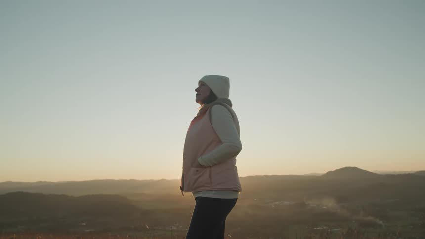 A woman with a hat and a vest standing with a landscape and the sky in the background
