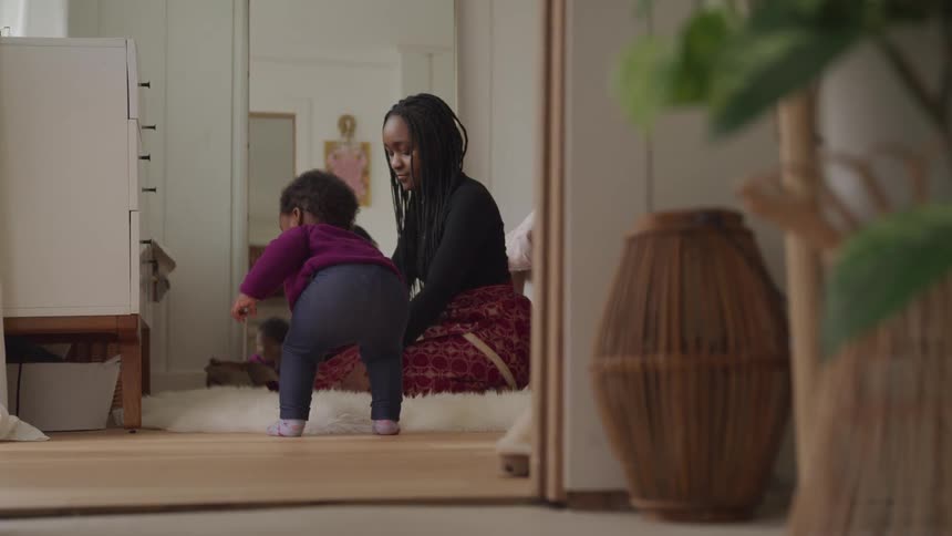 A little girl walking with a woman sitting on the floor with a mirror in the background