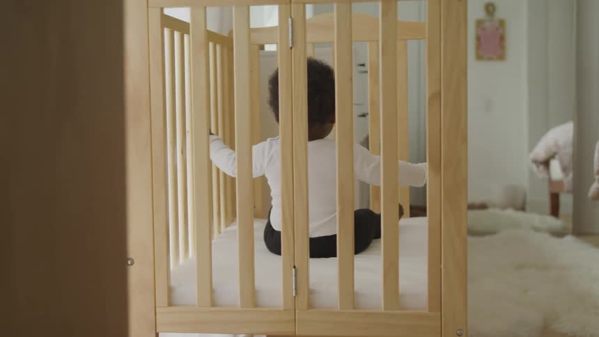 A baby boy stretches his arms out as he sits in a wooden crib on a furry white blanket