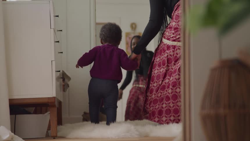 A woman holds a babys hand as they walk towards another woman in a home