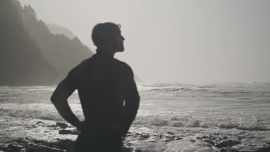 Silhouette of a young fit man wearing a wet suit stands pensively on the coast next to the ocean