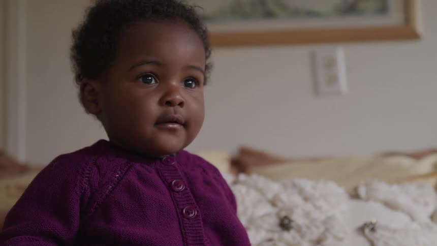 Black toddler girl sits on a bed and looks around