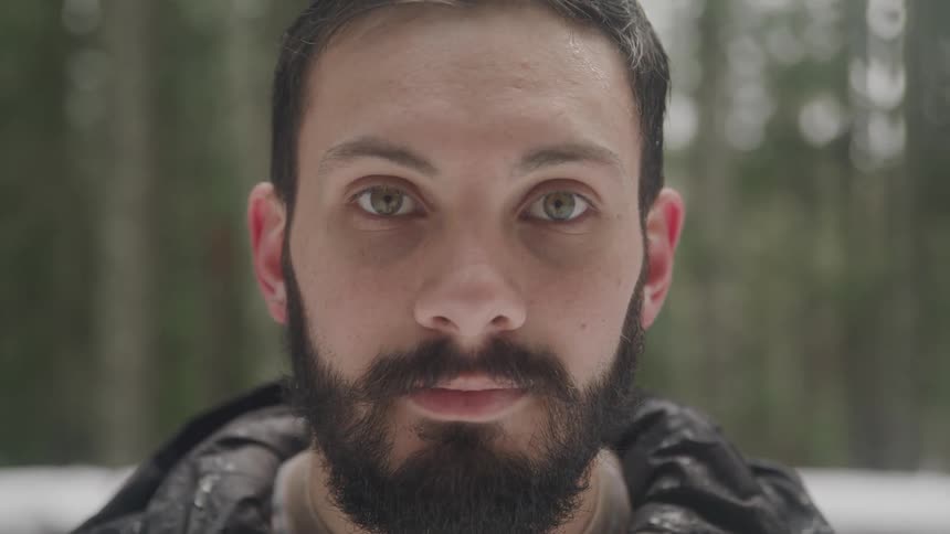 Man with a beard and hazel eyes raises his head and faces the camera with eye contact in the forest