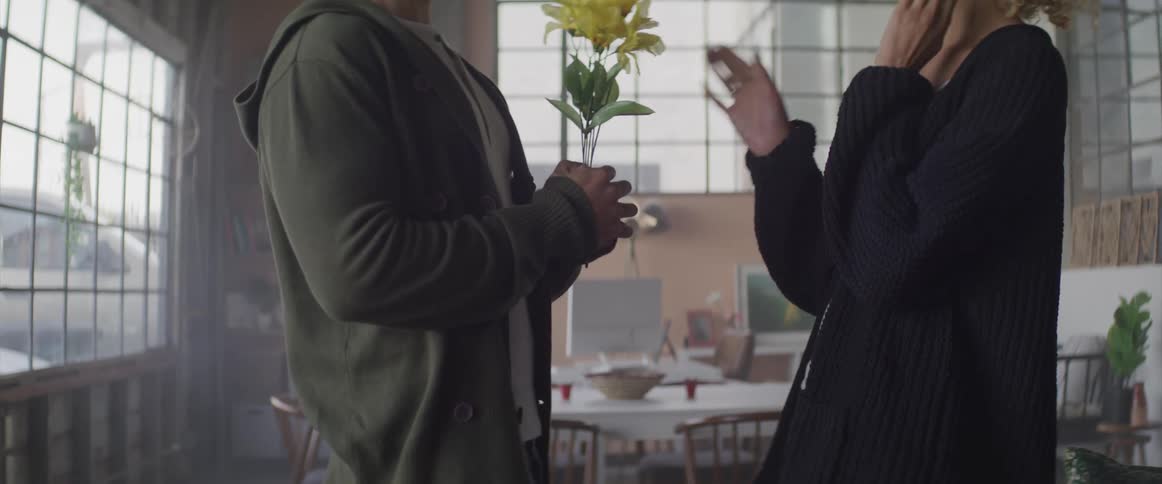 A man hides flowers behind his back and then pulls them around to surprise the woman standing in front of him