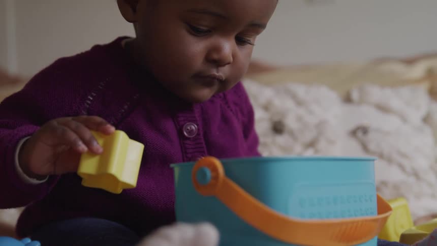 A baby sits in front of a toy bucket and holds a block