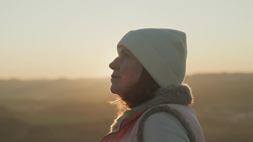 A woman is standing outdoors at dusk while wearing a hat